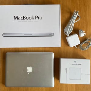 Könnte beinhalten: Ein silberner MacBook Pro Laptop mit der originalen weißen Verpackung, einem weißen 60W MagSafe Netzteil und einer separaten weißen Netzteilbox. Der Laptop hat eine metallische Oberfläche mit einem Apple-Logo auf dem Deckel und ein Stromkabel.