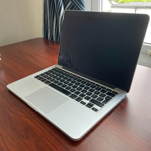 Può includere: Un computer portatile color argento con tastiera nera e un ampio touchpad, caratterizzato da uno schermo scuro. Un cavo di alimentazione bianco avvolto in un cerchio si trova a sinistra del dispositivo, poggiato su una superficie in legno scuro.
