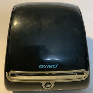 Op de afbeelding: Een zwarte Dymo LabelWriter 4XL labelprinter met een glanzende afwerking. Het apparaat heeft een lichtgrijze horizontale sleuf voor labeluitvoer, een kleine ronde knop onder de sleuf en de merknaam Dymo in het lichtblauw op de voorzijde.