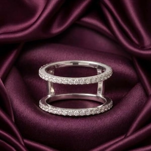 May include: A double band ring crafted from polished silver-toned metal, featuring two parallel bands connected by vertical bars. Both bands are encrusted with a continuous row of small, clear, sparkling stones that catch the light.