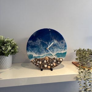 May include: A circular wall clock featuring a resin art design of an ocean shoreline with deep blue water, white foam waves, and a sandy beach base embedded with various small seashells and stones, complete with simple white clock hands.