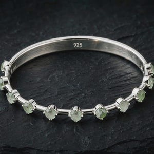 Könnte beinhalten: Ein silberner Ring mit einem dünnen Band und einer Reihe kleiner, runder, blassgrüner Edelsteine entlang der Außenkante. Die Innenseite des Rings ist deutlich mit der Zahl 925 geprägt, was auf die Zusammensetzung aus Sterlingsilber hinweist.