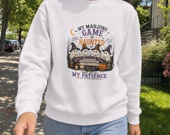 May include: A white crewneck sweatshirt featuring a graphic print of four ghosts wearing witch hats playing a mahjong game. The text reads: My Mahjong Game Is Haunted But So Is My Patience. Play At Your Own Risk. Includes a pumpkin and skull.