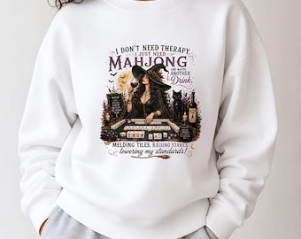 Op de afbeelding: Een wit sweatshirt met ronde hals en een grafische print van een heks die mahjong speelt met een zwarte kat, een wijnglas en een fles. De tekst luidt: I DON'T NEED THERAPY, I JUST NEED MAHJONG AND MAYBE ANOTHER DRINK. MELDING TILES, RAISING STAKES, lowering my standards!