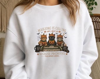 May include: A white crewneck sweatshirt featuring a graphic of three cats wearing pumpkin hats playing mahjong. The text reads: MAHJONG IS CALLING AND I MUST GO BECAUSE PRIORITIES. TILES BEFORE CHORES. Includes small bats, a spiderweb, and a coffee mug.