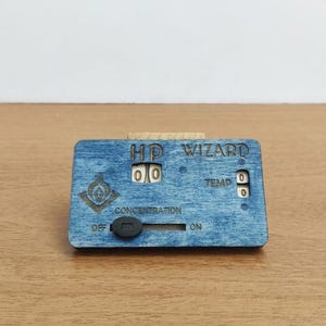 Może przedstawiać: Niebieski, prostokątny drewniany licznik do gry z wygrawerowanym napisem HP WIZARD, TEMP oraz CONCENTRATION. Zawiera dwa zestawy obrotowych kółek z cyframi, czarny przełącznik suwakowy z oznaczeniami OFF i ON oraz małą grafikę w kształcie płomienia.