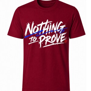 Op de afbeelding: Een bordeauxrood t-shirt met korte mouwen en de witte tekst "NOTHING TO PROVE" in een penseelstreeklettertype. Een dunne blauwe horizontale lijn loopt door de tekst en rechts staat een klein rood symbool voor een geregistreerd handelsmerk.