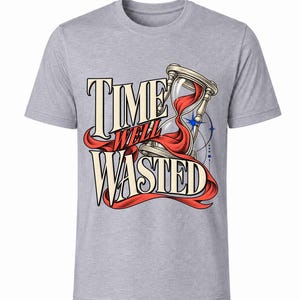 Op de afbeelding: Een grijs gemêleerd T-shirt met korte mouwen en een grafische illustratie van een zandloper waar rood zand doorheen stroomt. De tekst "TIME WELL WASTED" staat in een vet, gestileerd lettertype in het midden, vergezeld door een blauwe ster.