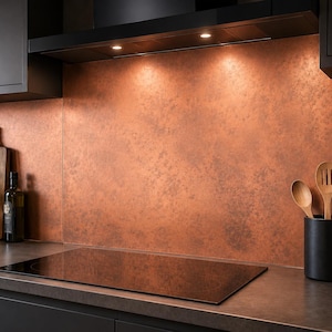 May include: A kitchen splashback featuring a textured copper-coloured finish behind a black glass induction hob. The area includes black cabinetry, a wooden chopping board, a glass bottle, and a black utensil holder with wooden spoons.