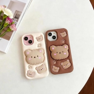 May include: Two smartphone cases featuring raised 3D bear designs. One case is cream-colored with light brown bear faces and paw prints, while the other is dark brown with matching bear faces and paw prints. Both have cutouts for camera lenses.
