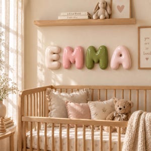 May include: Wall-mounted balloon-style letters spelling EMMA in cream, pink, and olive green. The letters are displayed above a wooden crib containing two decorative pillows, a beige teddy bear, and a cream-colored knit blanket.