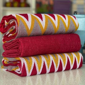 May include: A stack of three folded textiles. The top and bottom pieces feature a repeating chevron pattern in red, white, yellow, and grey. The middle piece is a solid, textured deep red fabric. Each textile has a woven, durable appearance.