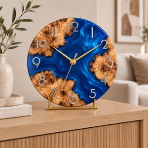 May include: A round table clock featuring a natural wood burl edge and a central blue epoxy resin river design. The clock face displays silver-colored numerals from 1 to 12 and thin gold-toned hands, supported by a gold metal stand on a flat surface.