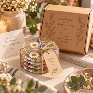 Puede incluir: Una pila de cuatro posavasos de resina transparente con margaritas blancas y flores azules prensadas, atados con cuerda y una etiqueta que dice A Brighter Home. Al lado hay una caja de cartón marrón con el texto Good Things for a Brighter Home.