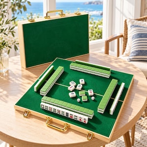 Op de afbeelding: Een Mahjong-spelset met een houten koffer met een groene vilten binnen- en buitenkant. De set bevat rechthoekige tegels met groene achterkanten en witte voorkanten gemarkeerd met zwarte en rode symbolen, samen met twee kleine witte dobbelstenen.
