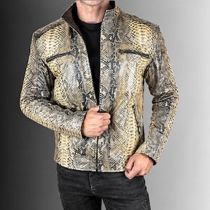 May include: A jacket featuring a snakeskin pattern in shades of tan, beige, and black. The garment has a stand-up collar, a full-length front zip, and two horizontal zipped chest pockets. The interior lining is dark brown, visible at the collar.