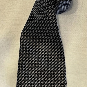 May include: A necktie featuring a repeating geometric pattern of small, light grey rectangular shapes arranged in a grid against a dark black background. The tie is partially coiled at the top, showing the consistent pattern across the fabric surface.