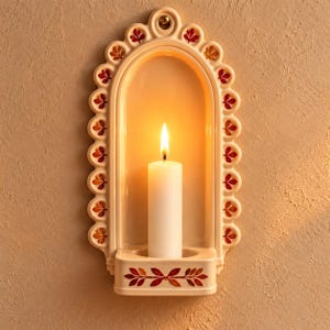 Puede incluir: Un aplique de pared de cerámica color crema con diseño arqueado y borde festoneado con pequeños motivos de hojas rojas y naranjas. El aplique tiene una plataforma central que sostiene una vela blanca con una llama brillante y cálida.