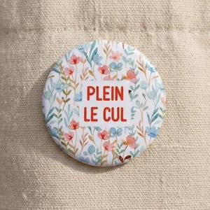 Peut inclure: Un badge rond épinglé sur un sac en toile beige. Le badge présente un motif floral délicat avec des petites fleurs et feuilles rouges, bleues et vertes. Au centre, le texte PLEIN LE CUL est imprimé en lettres capitales rouges et grasses.