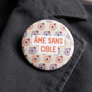Peut inclure: Un badge rond avec un motif répétitif d'yeux violets centrés dans des cibles circulaires orange et bleues sur fond crème. Le texte "ÂME SANS CIBLE" est imprimé en lettres orange sur un bloc rectangulaire blanc au centre du badge.