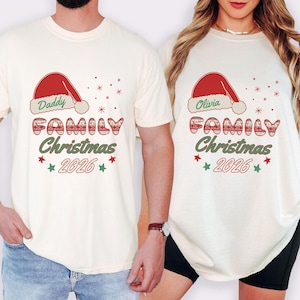 May include: Two cream-colored t-shirts featuring a red Santa hat graphic above the text "Daddy" or "Olivia" and the word "FAMILY" in a red patterned font. Below, the text "Christmas" and "2026" appear in green and red with small star accents.