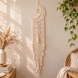 May include: A cream-colored macrame wall hanging featuring a crescent moon shape at the top with a smaller moon charm suspended inside. The design includes intricate knotted patterns, vertical fringe strands, and wooden beads throughout the piece.
