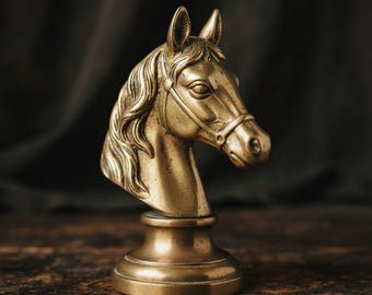 Puede incluir: Una escultura de cabeza de caballo de latón con una crin detallada, rasgos faciales definidos y una brida. La escultura está montada sobre una base circular de pedestal, mostrando un acabado dorado metálico con sutiles acentos de pátina oscura.