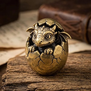 May include: A small, antique-style brass figurine depicting a baby dragon emerging from a cracked egg. The dragon features detailed scales, small wings, and horns, with its front claws resting on the jagged edge of the metallic eggshell.