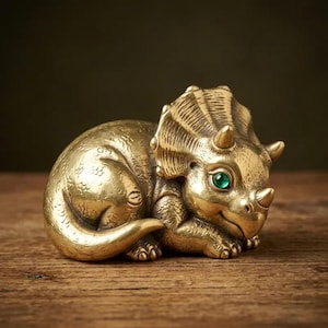 May include: A brass triceratops figurine featuring a textured, scale-like surface, a large frill, three horns, and a single bright green gemstone set into the eye socket. The dinosaur is depicted in a resting, curled position with its tail tucked.