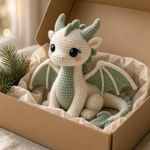 May include: A cream and sage green crocheted dragon plush toy with large black eyes and pink cheeks, sitting inside an open cardboard gift box lined with tissue paper, accompanied by a small sprig of green pine needles tucked into the corner.
