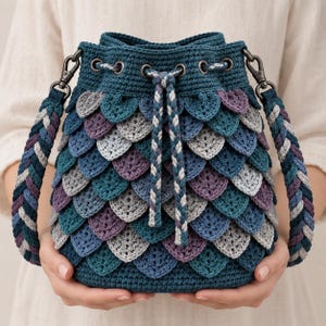 Può includere: Una borsa a secchiello con base all'uncinetto color verde ottanio scuro, caratterizzata da strati sovrapposti a forma di squame nei toni del verde ottanio, viola e grigio chiaro. Include una chiusura a coulisse intrecciata e una tracolla spessa.