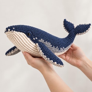 Puede incluir: Un juguete de ballena tejido a ganchillo con el cuerpo superior azul marino oscuro y la parte inferior de color crema con textura acanalada. La ballena tiene una aleta caudal ancha, dos aletas laterales y un pequeño ojo negro, todo tejido.
