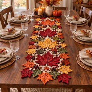 Peut inclure: Un chemin de table en crochet orné d'un motif répétitif de feuilles d'érable dans des tons de rouge, orange, jaune et vert, agrémenté de petits glands crochetés. Le chemin est centré sur une table à manger en bois avec des assiettes.