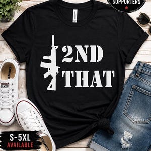 Peut inclure: Un t-shirt noir à manches courtes avec une impression graphique blanche représentant la silhouette d'un fusil à côté du texte 2ND THAT. Le t-shirt a un col rond et est présenté à plat avec des baskets blanches et un jean bleu visibles.