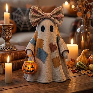 Pode incluir: Uma figura de fantasma de tecido com um laço de estampa floral na cabeça e quadrados de patchwork coloridos no corpo. O fantasma segura um pequeno balde de abóbora laranja. Está rodeado por velas acesas, pequenas abóboras e folhas de outono.