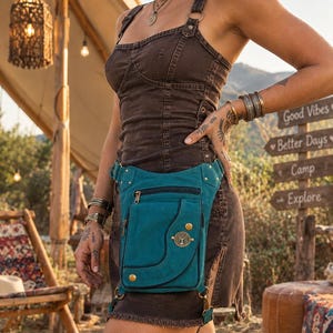 Puede incluir: Una bolsa de pierna de lona color verde azulado con múltiples compartimentos con cremallera, herrajes en tono latón y un emblema metálico circular que representa un árbol. La bolsa incluye correas ajustables y un bolsillo con solapa.