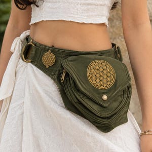 May include: An olive green utility belt featuring a large triangular pocket with a gold-toned flower of life embroidery, a smaller circular metal emblem with matching geometric patterns, a side zip, and a metallic snap button closure.
