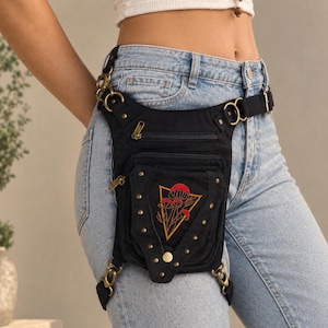 Puede incluir: Una bolsa de pierna de lona negra con un diseño bordado de setas rojas dentro de un triángulo dorado. La bolsa incluye múltiples compartimentos con cremallera, herrajes de metal en tono latón antiguo, tachuelas y correas ajustables para la cintura y el muslo.