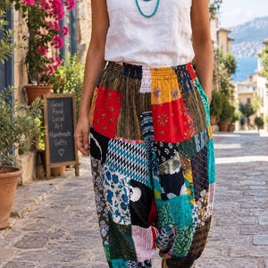 May include: A white sleeveless top with a turquoise beaded necklace and loose-fitting patchwork trousers featuring a variety of colorful floral, geometric, and striped patterns in shades of red, yellow, blue, green, black, and white.