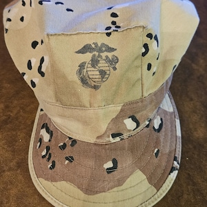 Puede incluir: Una gorra de patrulla con camuflaje desértico y un patrón de manchas en tonos beige, marrón y negro. El panel frontal muestra un emblema bordado en marrón oscuro de un águila, un globo terráqueo y un ancla sobre la visera a juego.