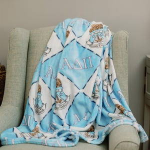 May include: A soft throw blanket featuring a repeating diamond pattern in light blue and white. The design includes the Greek letters AΔΠ and a crest emblem with a lion and anchor motif, printed across the fabric in a consistent, repeating layout.