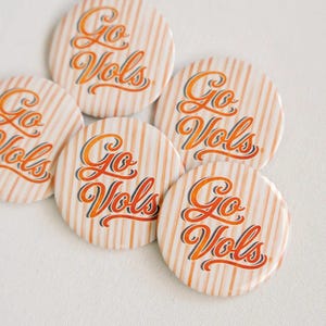 May include: Five circular pinback buttons featuring a white and orange vertical striped pattern. Each button displays the text Go Vols in a stylized orange cursive script with a dark blue outline, centered on the striped background.