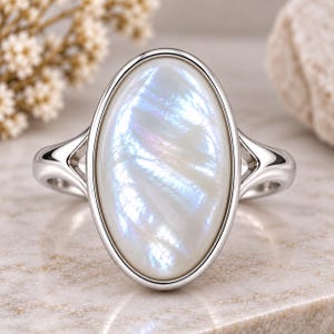 May include: A silver-toned ring featuring a large, oval-shaped, iridescent white mother-of-pearl cabochon set in a smooth metal bezel. The band has a split-shank design that connects to the central setting, showcasing a polished metallic finish.
