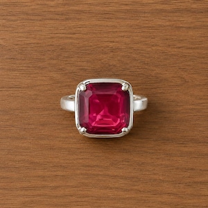May include: A silver ring featuring a large, square-cut, deep red gemstone set in a four-prong mounting. The band is smooth and polished, showcasing a clean, metallic finish that contrasts with the vibrant, faceted red stone at the center.
