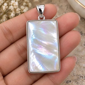 May include: A rectangular pendant featuring a smooth, iridescent white mother-of-pearl inlay set within a polished silver-toned metal frame. The surface of the stone displays subtle light-reflecting swirls of blue and pink hues.