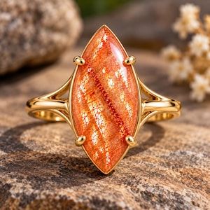Puede incluir: Un anillo de oro con una piedra de sol de corte marquesa en un tono naranja vibrante con inclusiones brillantes. La gema está asegurada por cuatro garras sobre una banda de oro pulido que se divide en dos segmentos finos en la montura.