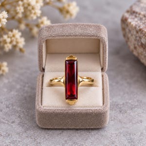 May include: A gold ring featuring a vertical, rectangular, deep red gemstone set in a polished gold band. The ring is displayed inside a beige, velvet-lined jewelry box, showcasing the contrast between the vibrant red stone and the gold metal.