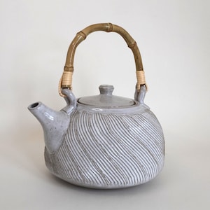 May include: A light grey ceramic teapot with a textured, diagonal ribbed pattern on the body. The teapot features a matching lid and a curved spout. It is equipped with a tall, arched handle made of natural bamboo, secured with light-coloured woven rattan.