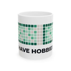 May include: A white ceramic mug featuring a graphic of green and light gray square tiles arranged in a grid pattern. Below the grid, the text "I HAVE HOBBIES" is printed in bold, black, sans-serif capital letters around the front of the mug.