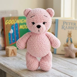 May include: A light pink crocheted teddy bear with a soft, plush texture. The bear features small round ears, black plastic eyes, and a stitched black nose and mouth. The yarn has a thick, velvety appearance, and the bear is standing upright.